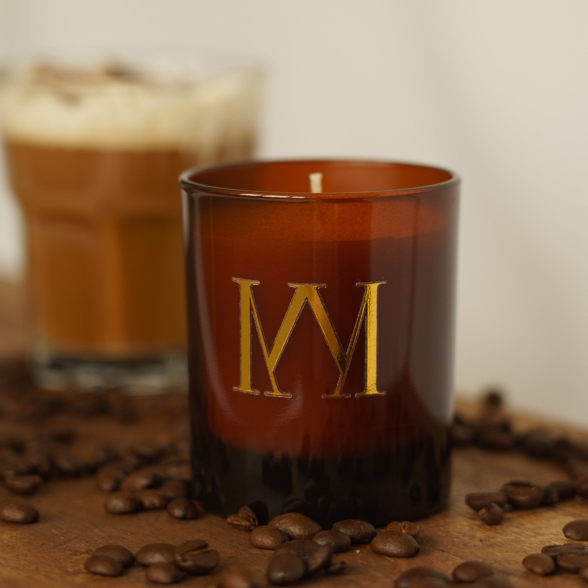 Candle with a gold 'M' logo on a wooden surface with coffee beans and a blurred cup in the background.