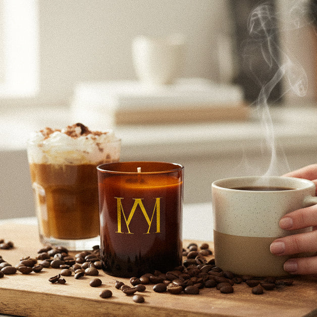 Candle with a logo, glass of coffee, and mug on a wooden surface with coffee beans.