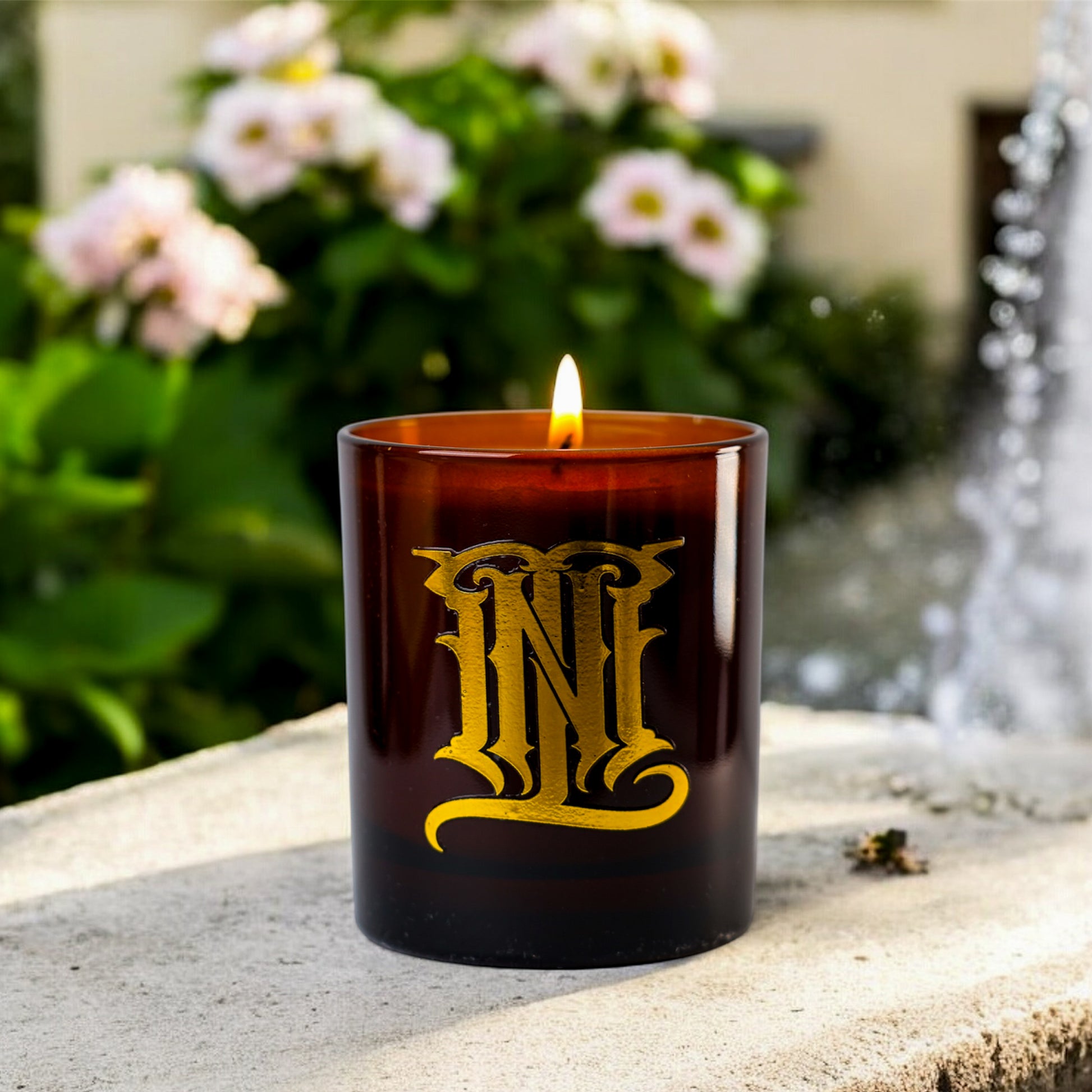 A CANDLE HOUSED IN AN AMBER JAR, WITH THE MONOGRAM 'MNL' ON A CONCRETE FOUNTAIN COUNTERTOP IN A GARDEN.
